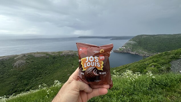 Un gâteau Jos Louis emballé tenu dans une main devant l'océan Atlantique et la verdure des côtes.