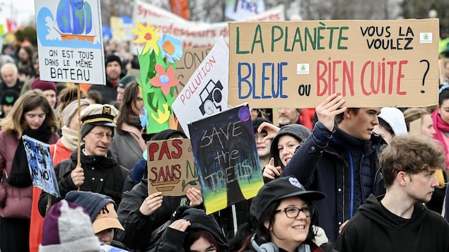 Des manifestants avec des pancartes aux messages variés, dont « La planète, vous la voulez bleue ou bien cuite? », « On est tous dans le même bateau » et « Sauvons les arbres ».