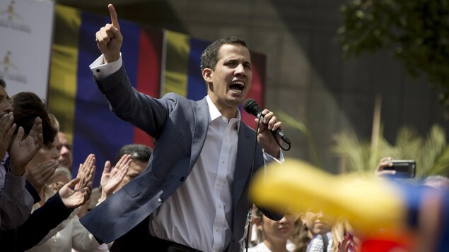 Juan Guaido parle dans un micro, sur une tribune. Des gens l'applaudissent.