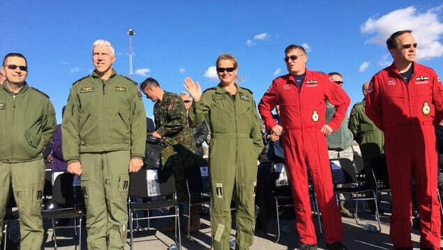 Julie Payette en combinaison de pilote salue la caméra, entourée d'autres pilotes. Julie Payette en combinaison de pilote salue la caméra, entourée d'autres pilotes.