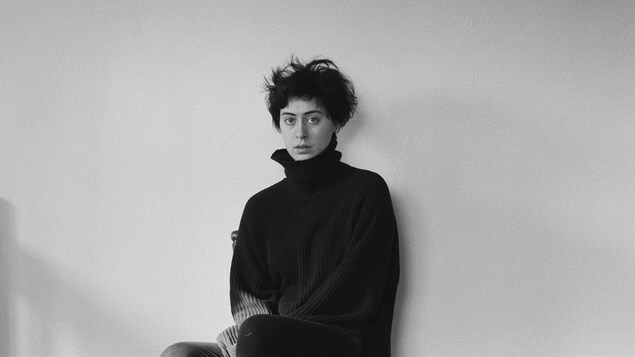 Portrait en noir et banc de Nina Schulman, assise sur une chaise, vêtue d'un chandail noir à col roulé relevé, cheveux courts en bataille.
