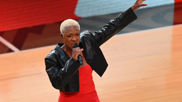 Una mujer, con un vestido rojo y una chaqueta de cuero negra, canta con un micrófono en el centro de una cancha de baloncesto.