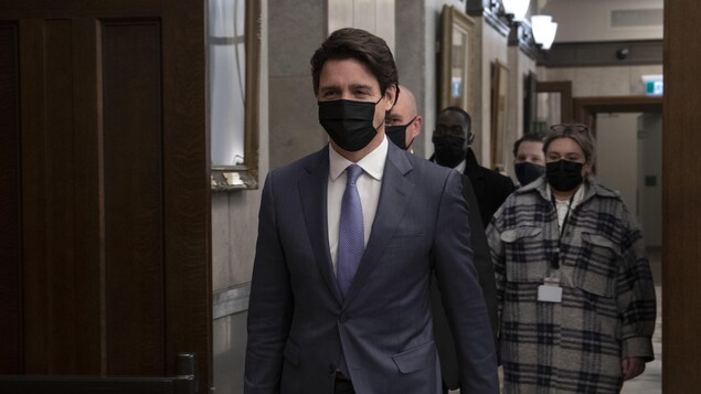 Justin Trudeau, his face masked, walks in a corridor followed by a few people, also masked.