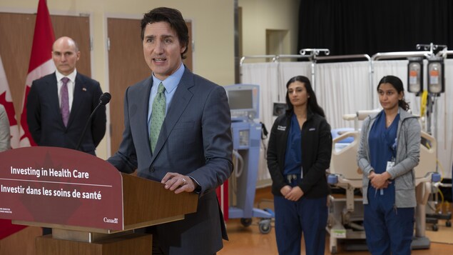 Justin Trudeau nagsasalita sa mikropono habang nasa background si Jean-Yves Duclos at mga babaeng health worker.