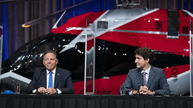 François Legault et Justin Trudeau sont assis à la table de presse devant un gros hélicoptère rouge. 