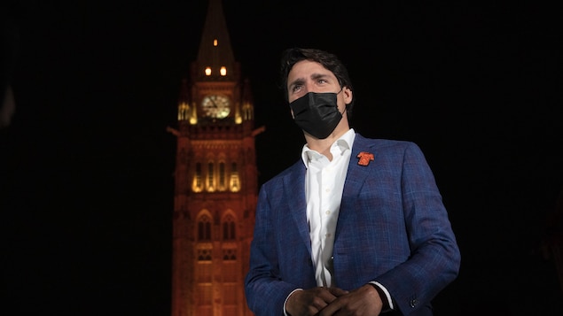 Justin Trudeau se tient debout, avec la Tour de la paix du Parlement canadien illuminée en arrière-plan.
