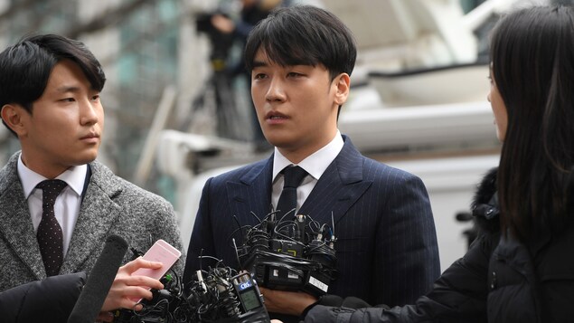 Le chanteur de K-pop, entouré de journalistes, parle face à des micros. 