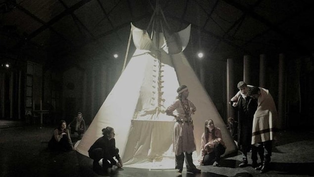 Des acteurs déguisés en Autochtones sont devant un tipi sur une scène.