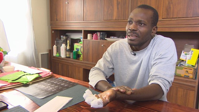 Un homme à la peau noire est assis à une table. Il porte un chandaille gris et montre sa main recouverte de bandage.