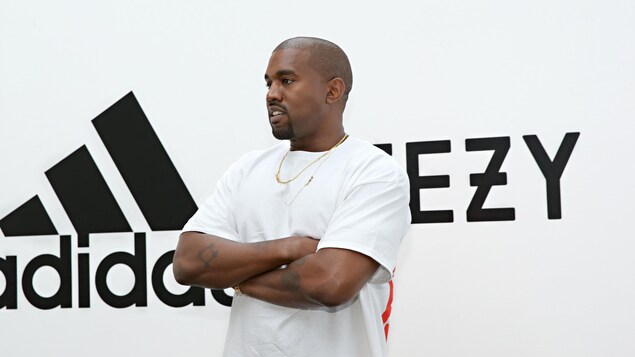 Un homme portant un t-shirt blanc pose les bras croisés devant un mur blanc affichant le logo d'Adidas et de yeezy. 