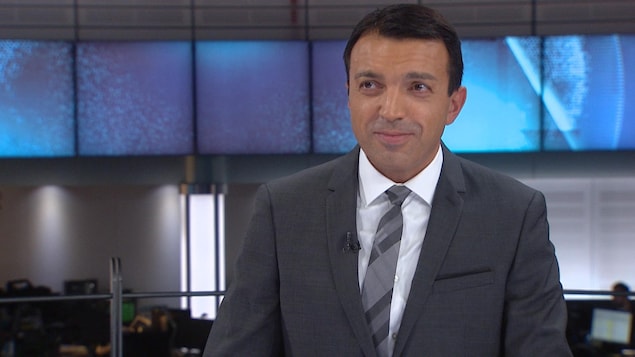 Un homme basané aux cheveux courts qui porte un veston, une blouse et une cravate regarde vers sa droite en souriant un peu.
