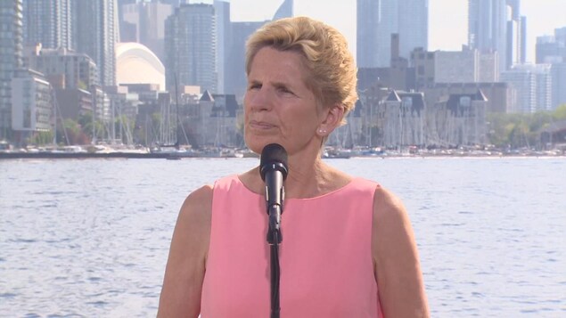 Photo de Kathleen Wynne portant un chemisier rose devant un micro, le lac Ontario en arrière-plan Photo de Kathleen Wynne portant un chemisier rose devant un micro, le lac Ontario en arrière-plan