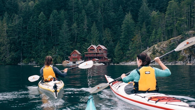 Deux femmes font du kayak en se dirigeant vers une habitation sur la côte.