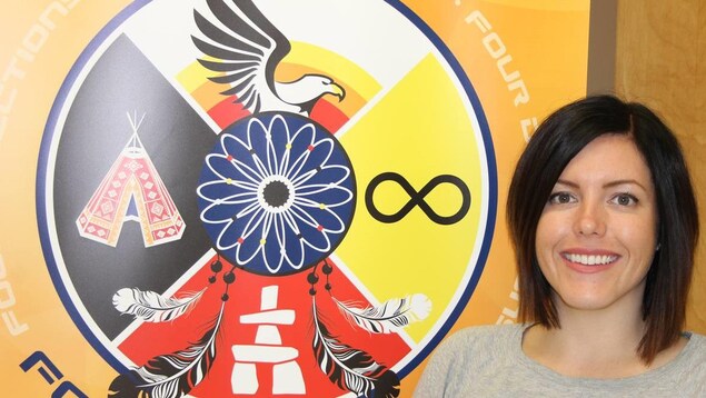 Une femme souriant à la caméra à côté d'un logo avec des symboles des Premières Nations, des Métis et des Inuits. Une femme souriant à la caméra à côté d'un logo avec des symboles des Premières Nations, des Métis et des Inuits.