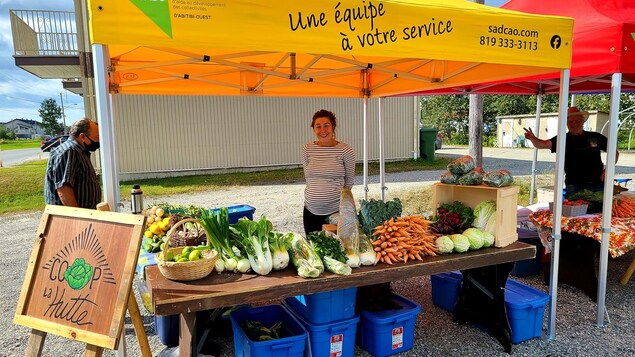 Une femme sourit derrière un kiosque avec une table remplie de légumes.