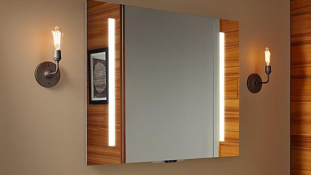 Une photo montrant un miroir installé dans une salle de bain. On peut apercevoir un petit appareil électronique dépassant du bas du miroir. Une photo montrant un miroir installé dans une salle de bain. On peut apercevoir un petit appareil électronique dépassant du bas du miroir.