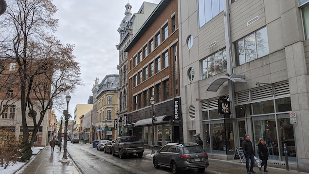 Des magasins, dont Laliberté, sur une rue commercial dans le quartier Saint-Roch à Québec. Quelques voitures sont stationnées et quelques passants marchent sur le trottoir.