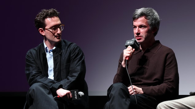 Les deux hommes sont assis dans une séance de questions, l'un d'eux s'exprimant dans un micro. 