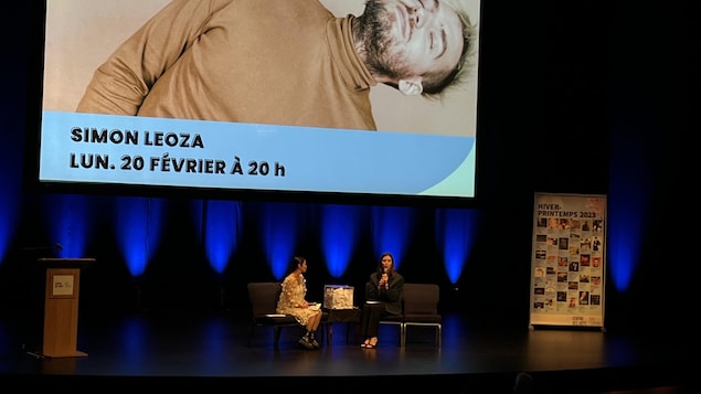 Deux animatrices sont assises sur des fauteuils sur la scène. Un écran présentant Simon Leoza se trouve derrière elles.