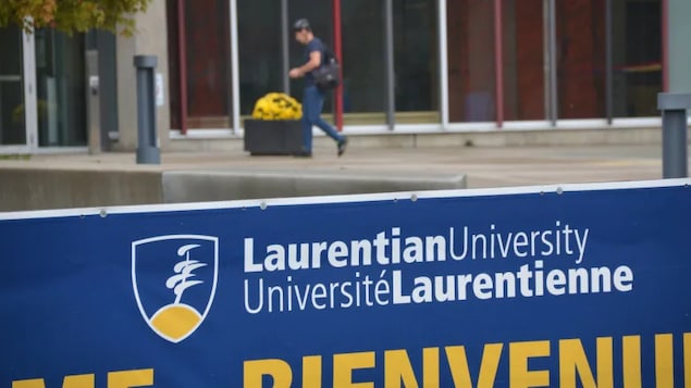 Un individu marche sur le campus de l'Université Laurentienne près d'un bâtiment, avec la pancarte de l'université en avant-plan.