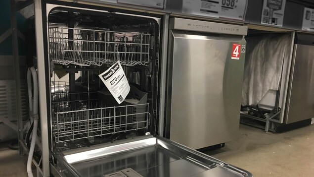 Des lave-vaisselle argentés, dont un qui est ouvert, sont présentés dans un magasin d'électroménagers. Des étiquettes et des papiers sont accrochés sur les machines.