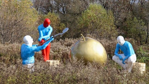 Trois grands personnages bleus sont autour d'une pomme dorée géante.