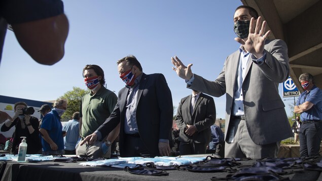 MM. Legault et Drouin sont derrière une table où sont étalés des masques jetables ou réutilisables pendant qu'un homme en veston fait signe aux curieux de garder leurs distances.