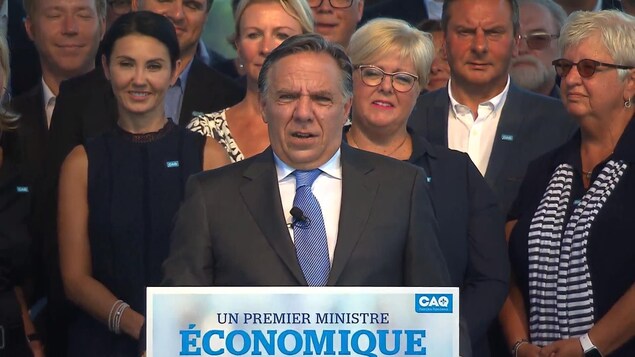 Un homme s'adresse à un auditoire derrière un lutrin sur lequel il est écrit: «Un premier ministre économique» avec le logo de la CAQ. D'autres personnes sont debout derrière lui.