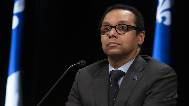 Lionel Carmant assis à la table de presse devant un drapeau du Québec affiche un air sérieux.