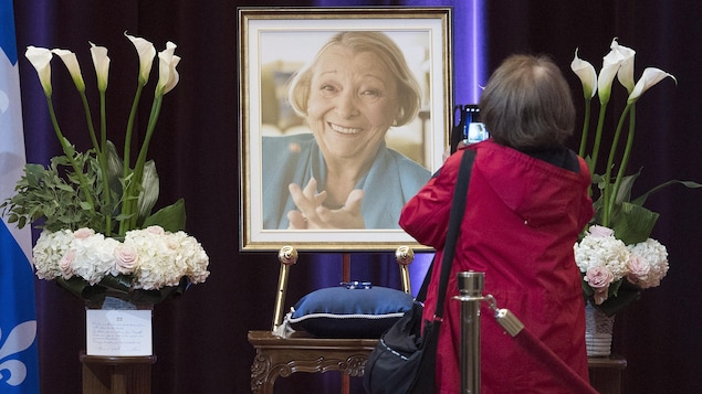 Une femme prend en photo un grand portrait de Lise Payette entouré de gerbes de fleurs.