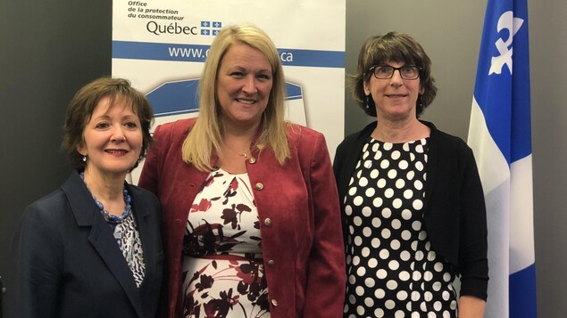 Trois femmes sourient à la caméra. Derrière elles, il y a une affiche de l'Office de la protection du consommateur. Trois femmes sourient à la caméra. Derrière elles, il y a une affiche de l'Office de la protection du consommateur.