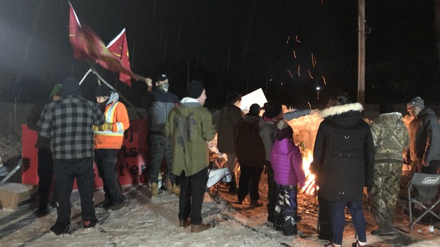 Une douzaine de personnes sont debout sur les rails enneigés et se réchauffent près d'un feu.