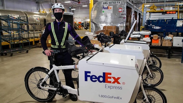 Un homme avec un masque et un casque tient un vélo de livraison dans un entrepôt.