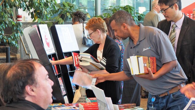 un homme prend un livre sur un étal, alors qu'il a déjà son autre main plein d'ouvrages. Derrière lui, des personnes regardent les livres présentés par les éditeurs, sur leurs stands. un homme prend un livre sur un étal, alors qu'il a déjà son autre main plein d'ouvrages. Derrière lui, des personnes regardent les livres présentés par les éditeurs, sur leurs stands.