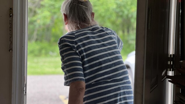 Un dame aux cheveux gris est de dos, dans un cadre de porte, et elle regarde vers l'extérieur.