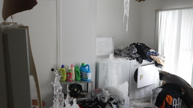 Une laveuse déborde de glace dans une salle de lavage où des vêtements sont empilés sur le sol et sur une sécheuse.