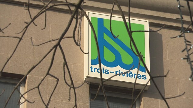 Le logo de la Ville de Trois-Rivières sur un bâtiment avec des branches dénudées de feuilles devant.