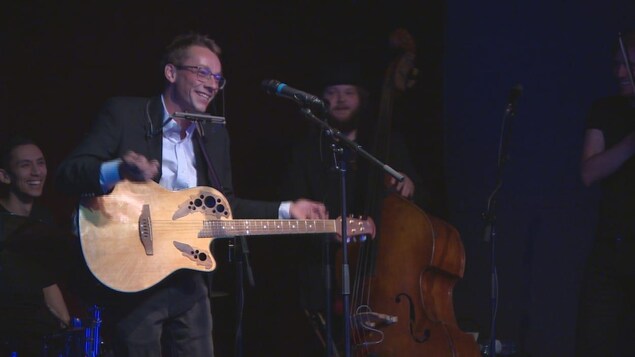 Lord Byrun sur scène devant un microphone, tenant une guitare et un harmonica.