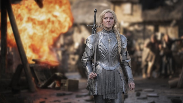Une femme portant une armure métallique marche sur un champ de bataille. On aperçoit au loin derrière elle une boule de feu accompagnant une déflagration.