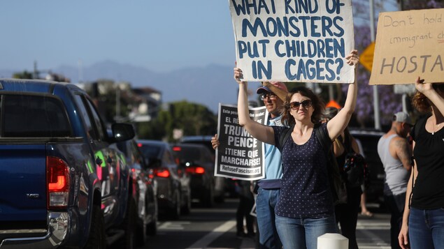 Des manifestants tiennent des affiches dénonçant la politique migratoire de l'administration Trump près d'une route où circulent des véhicules. Des manifestants tiennent des affiches dénonçant la politique migratoire de l'administration Trump près d'une route où circulent des véhicules.