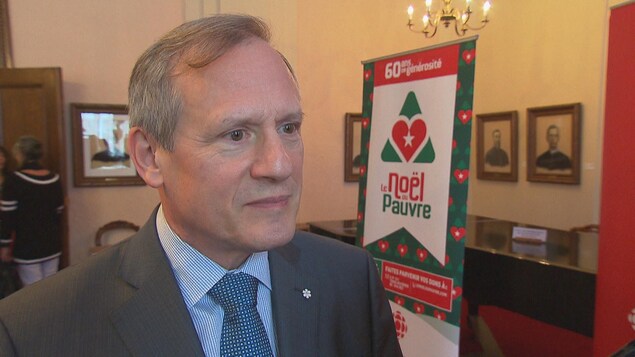 Un homme aux cheveux gris, devant une pancarte du Noël du Pauvre, écoute les questions des journalistes.