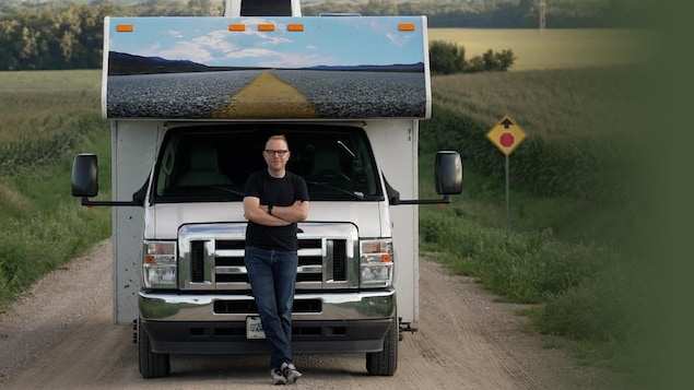 Un homme blanc est positionné debout devant un campeur, les bras croisés. Il regarde l'objectif de la caméra. Le campeur est stationné sur une route de campagne entre deux champs. 