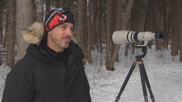 L'homme portant tuque et parka se tient debout à côté de son appareil photo à long objectif monté sur trépied dans une forêt.