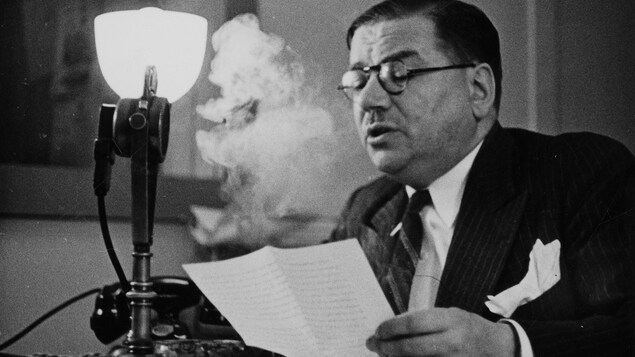 Dans un studio de radio, assis à un bureau, le journaliste Louis Francoeur parle au micro avec son texte dans la main. Une épaisse volute de fumée de sa cigarette monte au-dessus de la feuille.