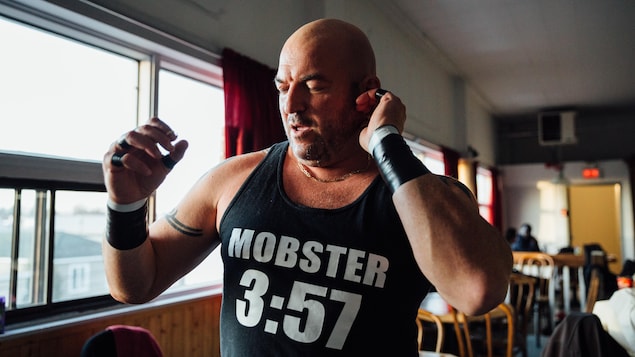 Un homme chauve porte une camisole sur laquelle est écrit «Mobster 3:57»