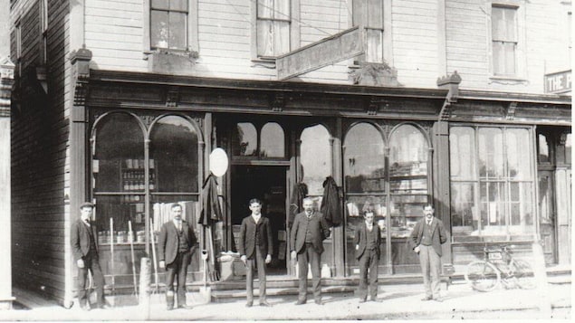 Six hommes se tiennent debout devant un magasin général dans les années 1900.