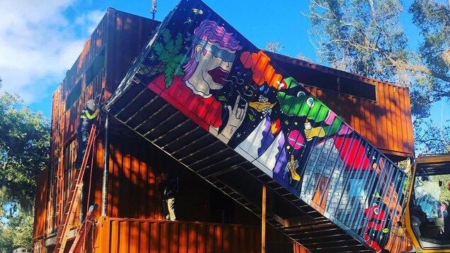 Un conteneur dont un des côtés affiche une murale en train d'être installé sur la maison à l'aide d'une grue.