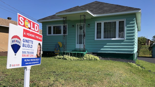 Une maison modeste avec une affiche d'une agente immobilière sur la pelouse avant.