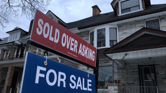 Une affiche de vente immobilière, sur laquelle on peut lire "Sold over asking" (vendu au-dessus du prix demandé), devant des maisons en rangée.