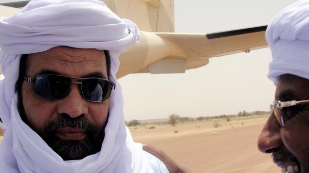 Un homme portant des lunettes de soleil, la barbe et un chèche blanc discute avec un autre homme vêtu de la même façon près d'un avion, au Mali. Un homme portant des lunettes de soleil, la barbe et un chèche blanc discute avec un autre homme vêtu de la même façon près d'un avion, au Mali.